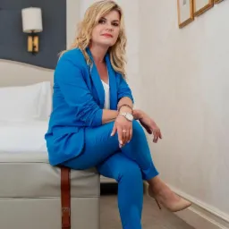 Kobieta w niebieskim garniturze siedzi na łóżku w pokoju hotelowym, spoglądając w obiektyw.