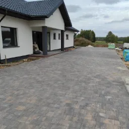 Nowoczesny dom z podjazdem wyłożonym szarą kostką brukową o prostokątnym kształcie, widoczne materiały budowlane w tle.