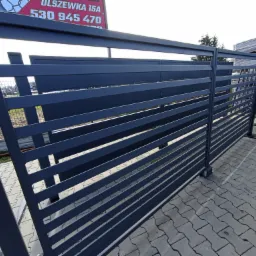 Nowoczesna, pozioma brama wjazdowa z szarych, metalowych profili, widoczna z boku na tle kostki brukowej i tablicy reklamowej.