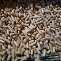 Jasne, cylindryczne pelety drzewne w dużym skupisku, część spada w dół, widoczna krawędź czarnej powierzchni pod spodem.