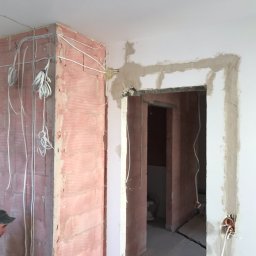 Crado - Przygotowanie do wykończenia wnętrza: ściana z instalacją elektryczną przed położeniem tynku, obrobione futryny drzwiowe, widoczny fragment osoby w czapce.