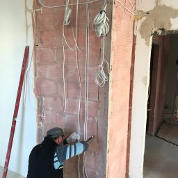 Crado - Instalacja elektryczna w trakcie montażu na ścianie z czerwonej cegły, widoczne przewody prowadzone przez pracownika w rękawicach, poziomica oparta o ścianę.