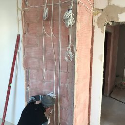 Crado - Instalacja elektryczna w trakcie montażu w nowo wznoszonym murze z czerwonej cegły, widoczne przewody, puszki, poziomica i pracownik w rękawicach.