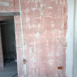 Crado - Surowa ściana z czerwonej cegły z widocznymi przewodami elektrycznymi w białych rurkach, wychodzącymi z puszek elektrycznych. Widoczne ślady po tynku. W tle widoczne kable elektryczne.