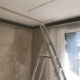 Crado - Pokój w trakcie wykańczania: surowe, otynkowane ściany, metalowe profile sufitowe, drabina aluminiowa i okno osłonięte folią malarską.