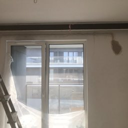 Crado - Wnętrze w trakcie prac wykończeniowych: ściana po tynkowaniu z widocznym metalowym profilem pod sufitem, okno zabezpieczone folią, drabina oparta o ścianę.