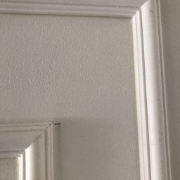 Crado - Fragment białej ściany z dekoracyjną sztukaterią i delikatną teksturą, widoczny mały defekt w rogu listwy.