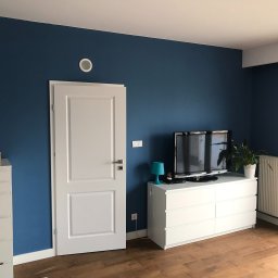 Crado - Pomieszczenie po malowaniu ścian na kolor ciemnoniebieski, z widocznymi białymi meblami, drzwiami i grzejnikiem, oraz drewnianą podłogą.