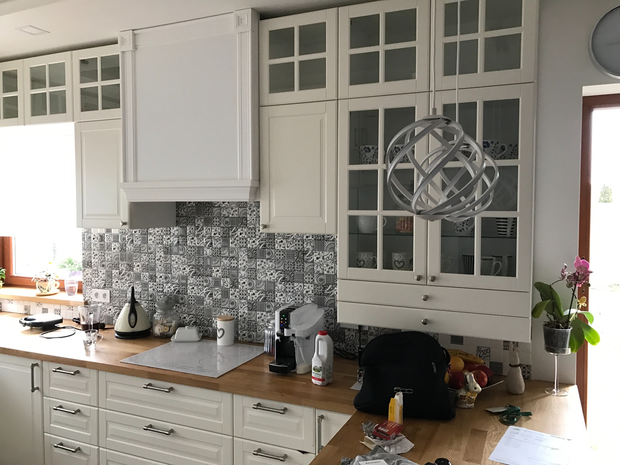 Jasna kuchnia z drewnianym blatem, białymi szafkami z przeszklonymi górnymi drzwiczkami, wzorzystą mozaiką na ścianie i nowoczesną lampą wiszącą.