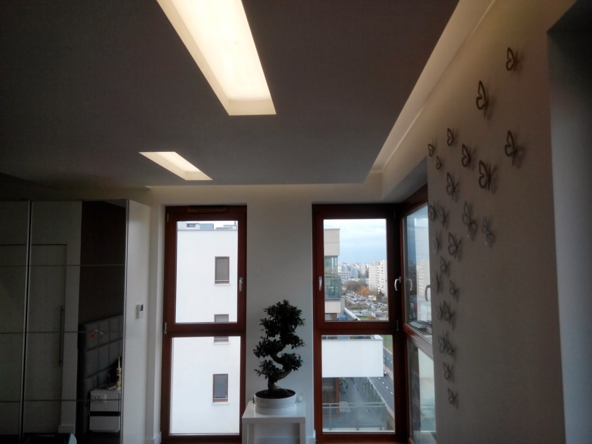 Jasne wnętrze mieszkania z oknami, dekoracją motyli na ścianie i podwieszanym sufitem z oświetleniem LED.