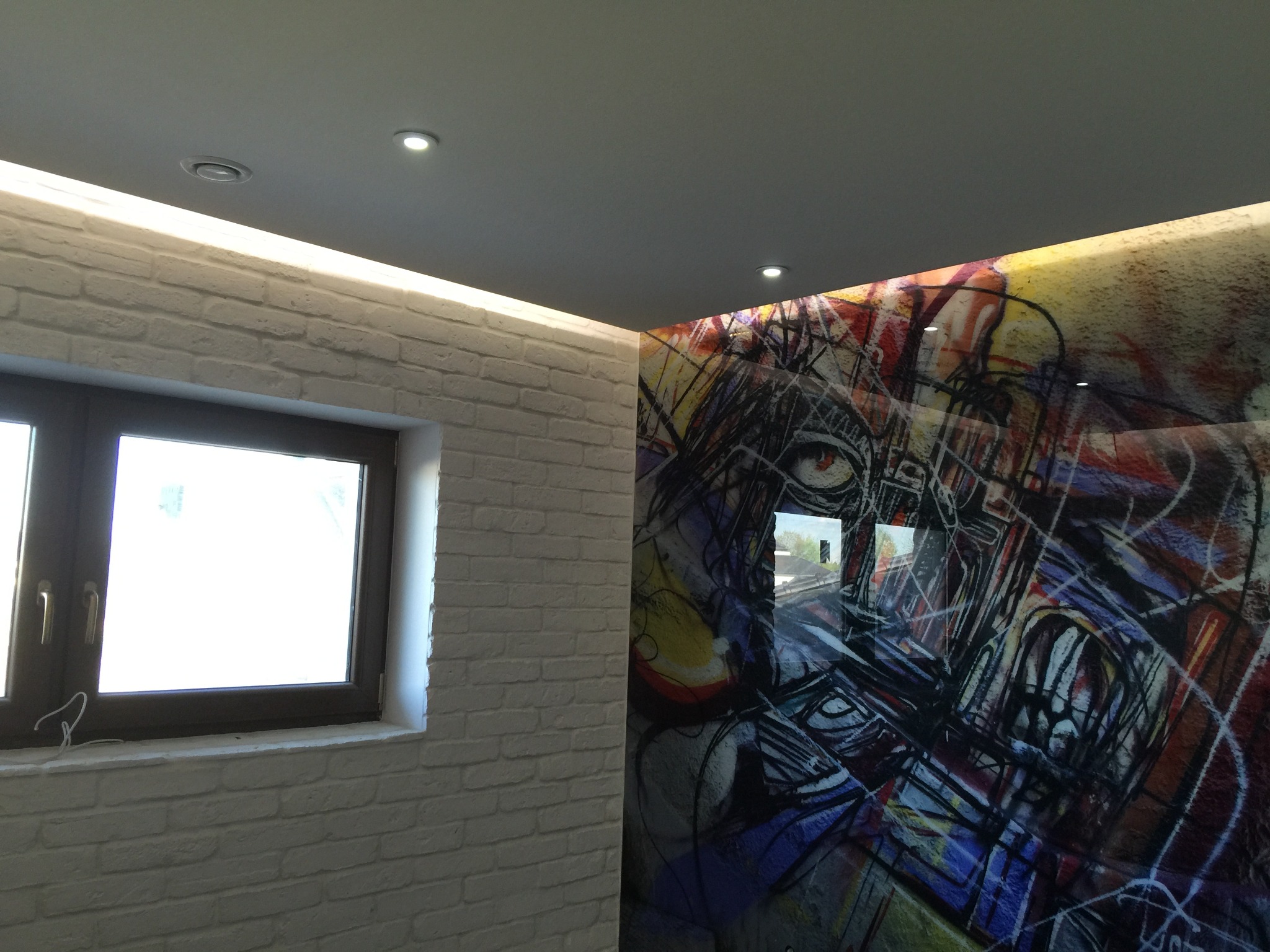 Wnętrze z białą cegłą na ścianie obok okna i szklanym panelem z abstrakcyjnym, kolorowym motywem grafitti na ścianie obok, oświetlone punktowymi lampami sufitowymi i listwą LED.