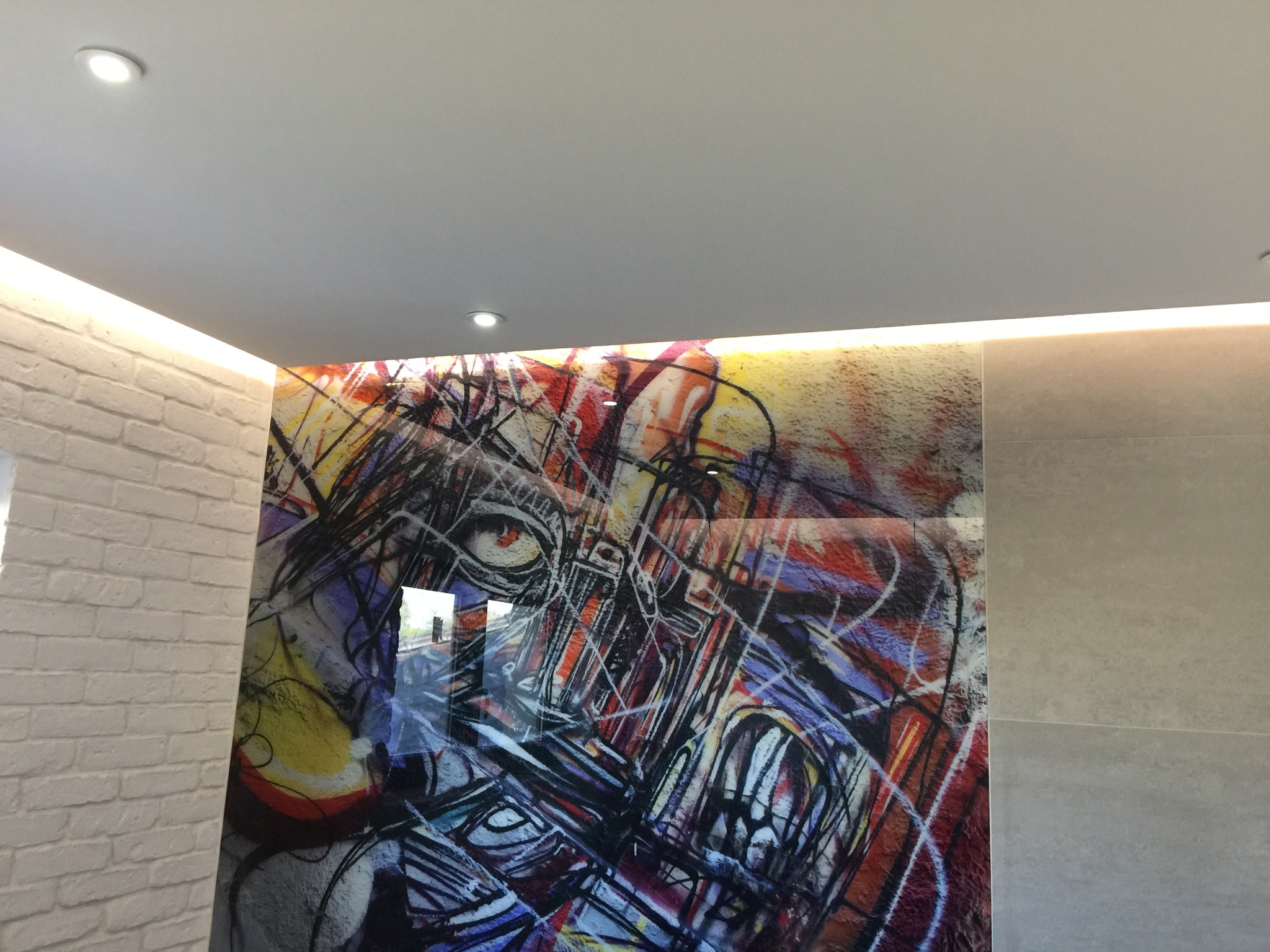 Ściana w łazience w Warszawie z dekoracyjną płytą szklaną z abstrakcyjnym, kolorowym wzorem graffiti, obok biała ściana z cegieł i szara ściana z płytek, oświetlenie LED pod sufitem.