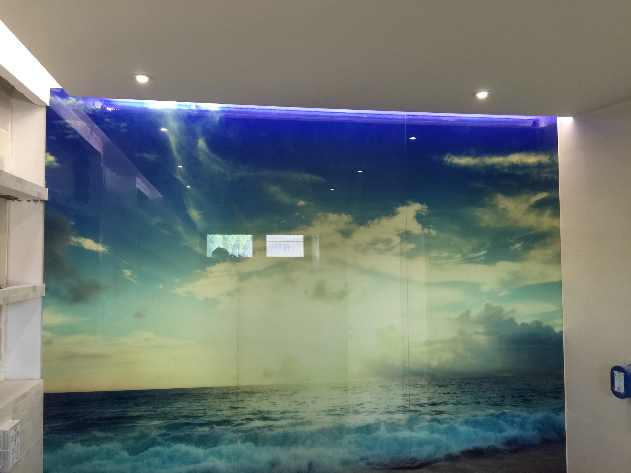 Ściana w łazience wykończona dużym panelem szklanym z nadrukiem motywu morskiego, imitującym widok oceanu i nieba, podświetlona od góry niebieskim światłem LED.