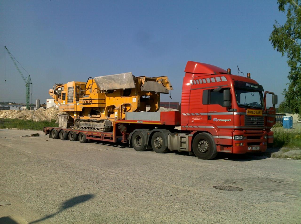 Czerwona ciężarówka MAN z naczepą niskopodwoziową przewożąca żółtą maszynę budowlaną EXTEC, widoczny żuraw budowlany w tle.