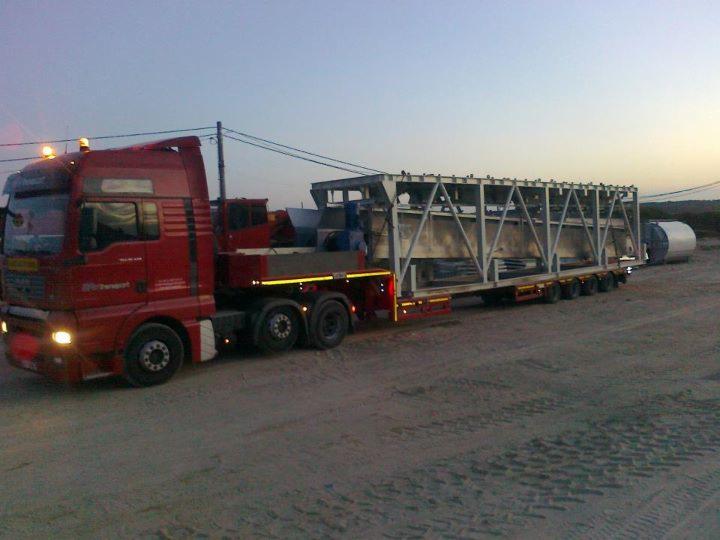 Czerwony ciągnik siodłowy z naczepą niskopodwoziową przewożący dużą, metalową konstrukcję szkieletową i element cylindryczny na piaszczystym terenie o zmierzchu.