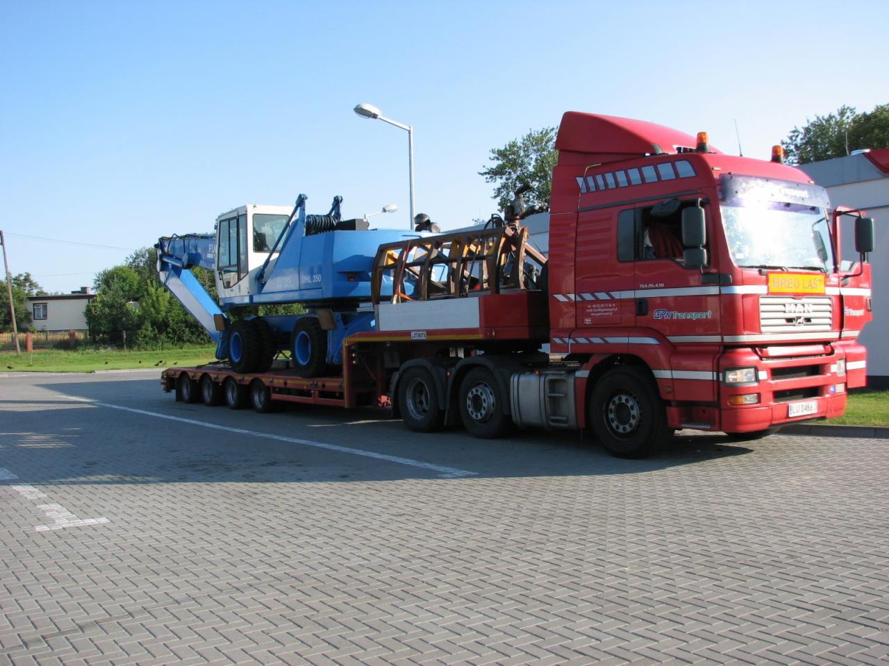 Czerwona ciężarówka MAN z naczepą niskopodwoziową przewożąca niebiesko-białą maszynę budowlaną typu WHL 350, widok z boku na placu wyłożonym kostką brukową.