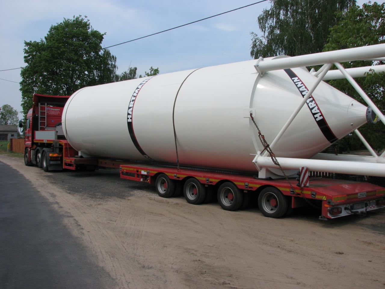 Czerwony ciągnik siodłowy z naczepą niskopodwoziową przewozi biały, cylindryczno-stożkowy zbiornik z napisem 'HARTMANN', zabezpieczony łańcuchami, na piaszczystej drodze wiejskiej.