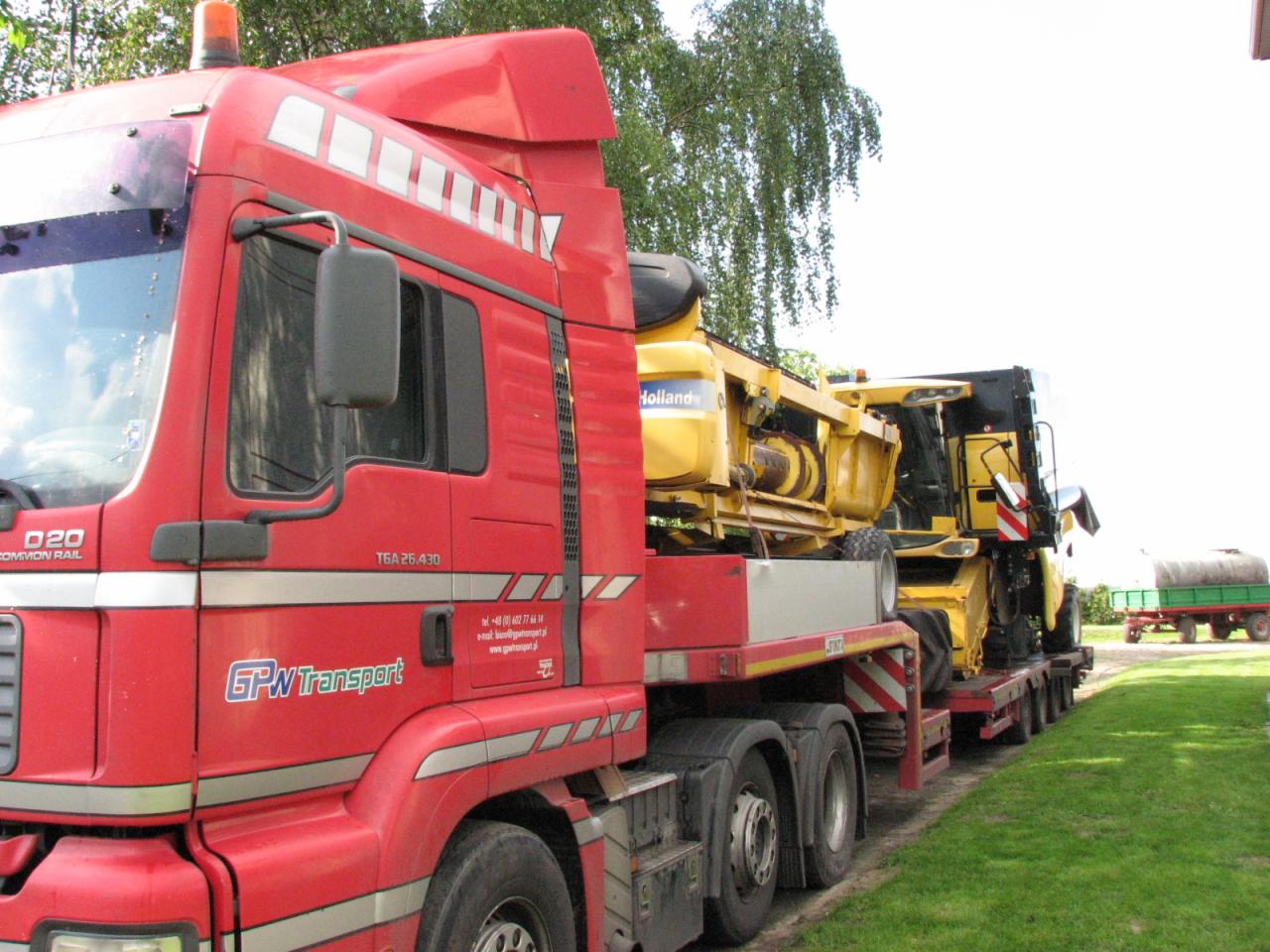 Czerwony ciągnik siodłowy GPW Transport z naczepą niskopodwoziową przewożący żółty kombajn rolniczy Holland na tle zieleni.