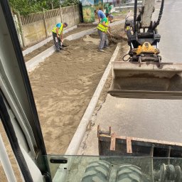 P.H.U Waldemar Ginter - Widok z kabiny koparki na robotników wyrównujących piasek pod chodnik, widoczne krawężniki i budynek w tle.