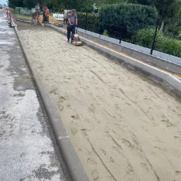 Robotnicy przygotowują podłoże pod chodnik z kostki, ubijając piasek zagęszczarką wibracyjną, widoczne nowe krawężniki, w tle drzewa.