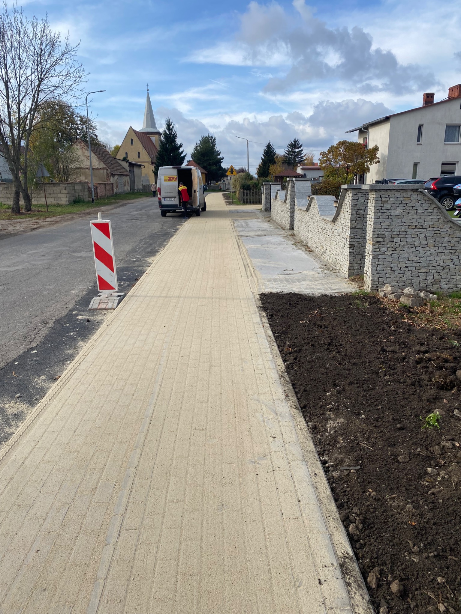 Nowo ułożona, jasna kostka brukowa chodnikowa z widocznym piaskiem fugującym, obok świeżo przekopana ziemia pod nasadzenia, w tle budynki mieszkalne i kościół, a także samochód dostawczy...