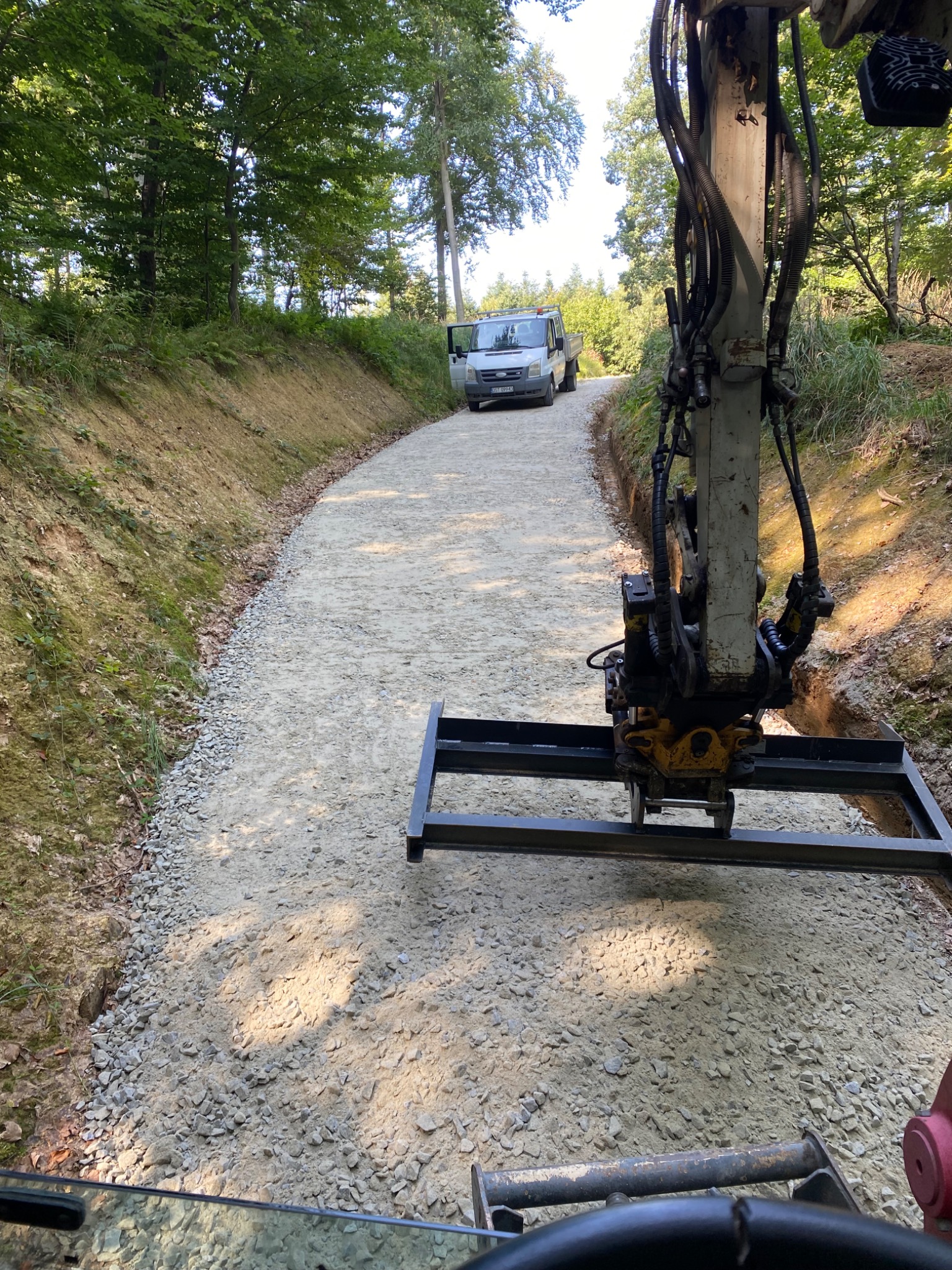 Niwelacja terenu pod drogę dojazdową w lesie, widoczny sprzęt budowlany i dostawczy bus na końcu drogi.