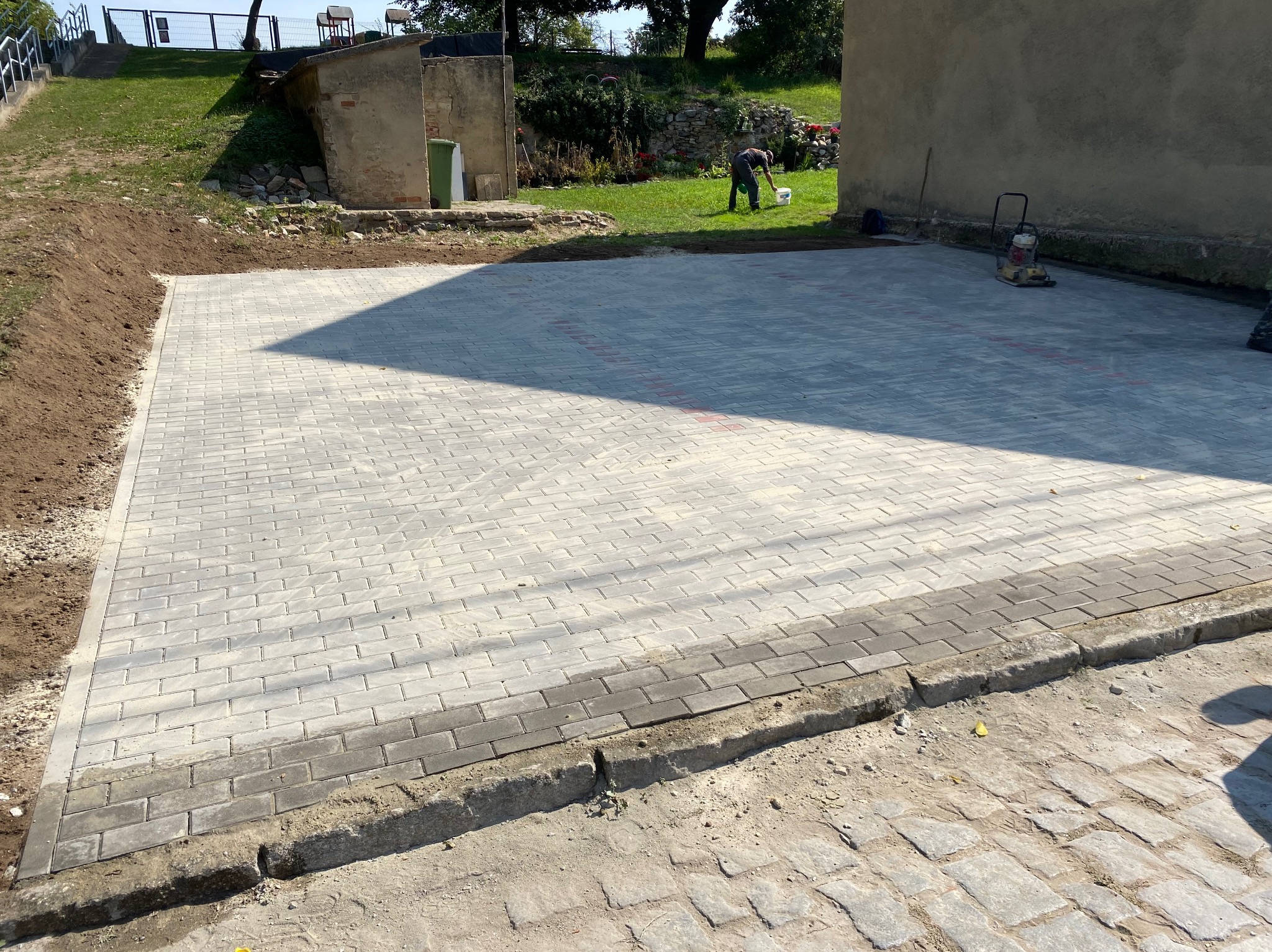 Nowo ułożona, jasnoszara kostka brukowa na przygotowanym podłożu, widoczny ubijak wibracyjny i pracownik w tle, krawężnik z ciemniejszej kostki oddziela nawierzchnię od ziemi.