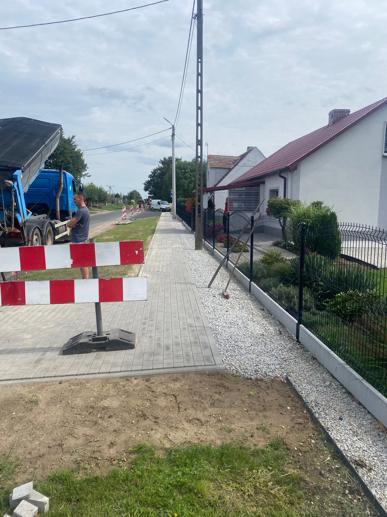 Nowo ułożona kostka brukowa chodnika z obrzeżem, widoczny ciężarowy samochód z wywrotką, biało-czerwone bariery ostrzegawcze oraz montaż ogrodzenia z metalowej siatki w tle zabudowa jednorodzinna.