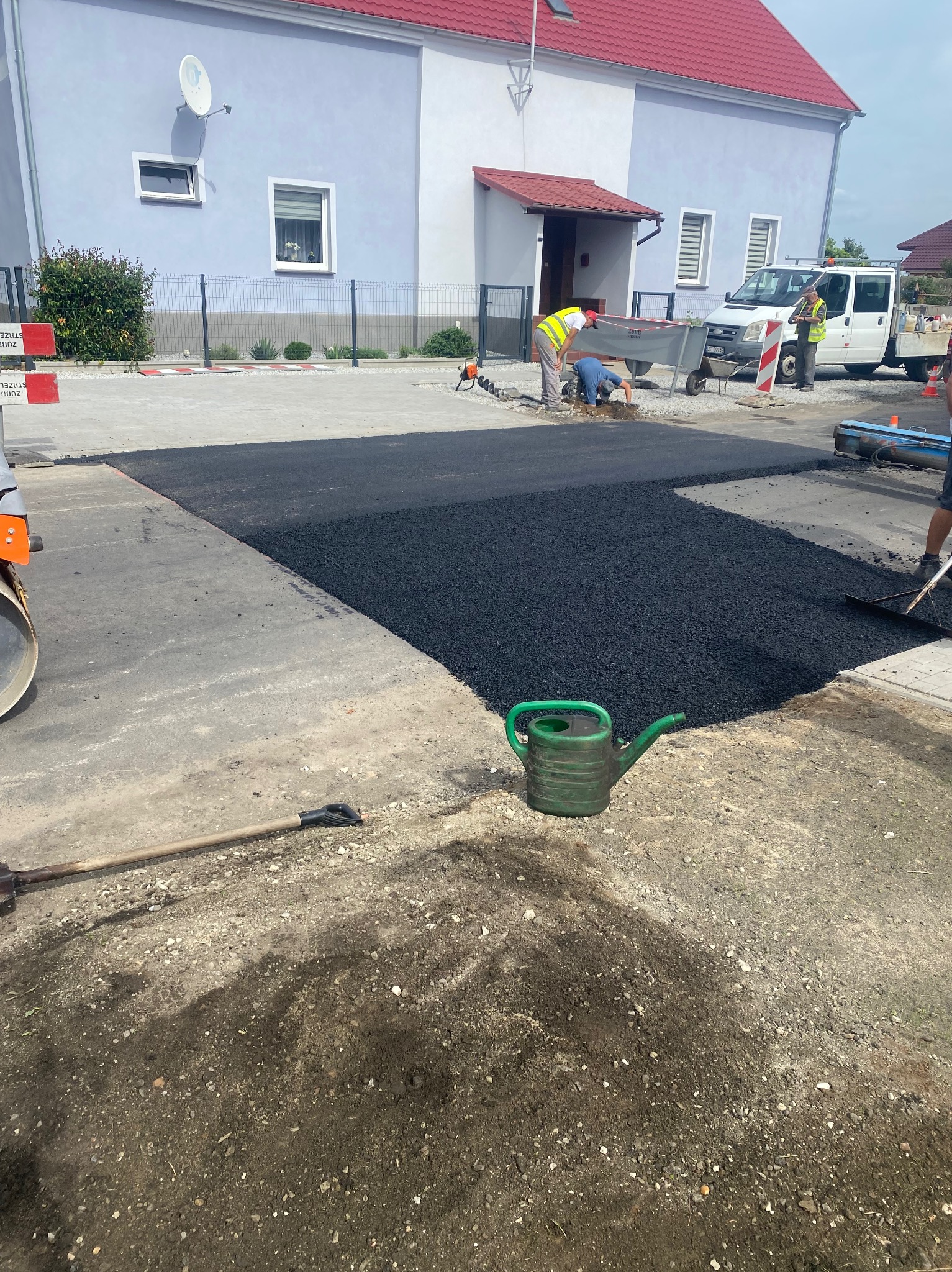 Prace drogowe: świeżo wylany asfalt na podjeździe przed domem, robotnicy wyrównują powierzchnię, w tle biały bus i dom z czerwonym dachem, na pierwszym planie zielona konewka.