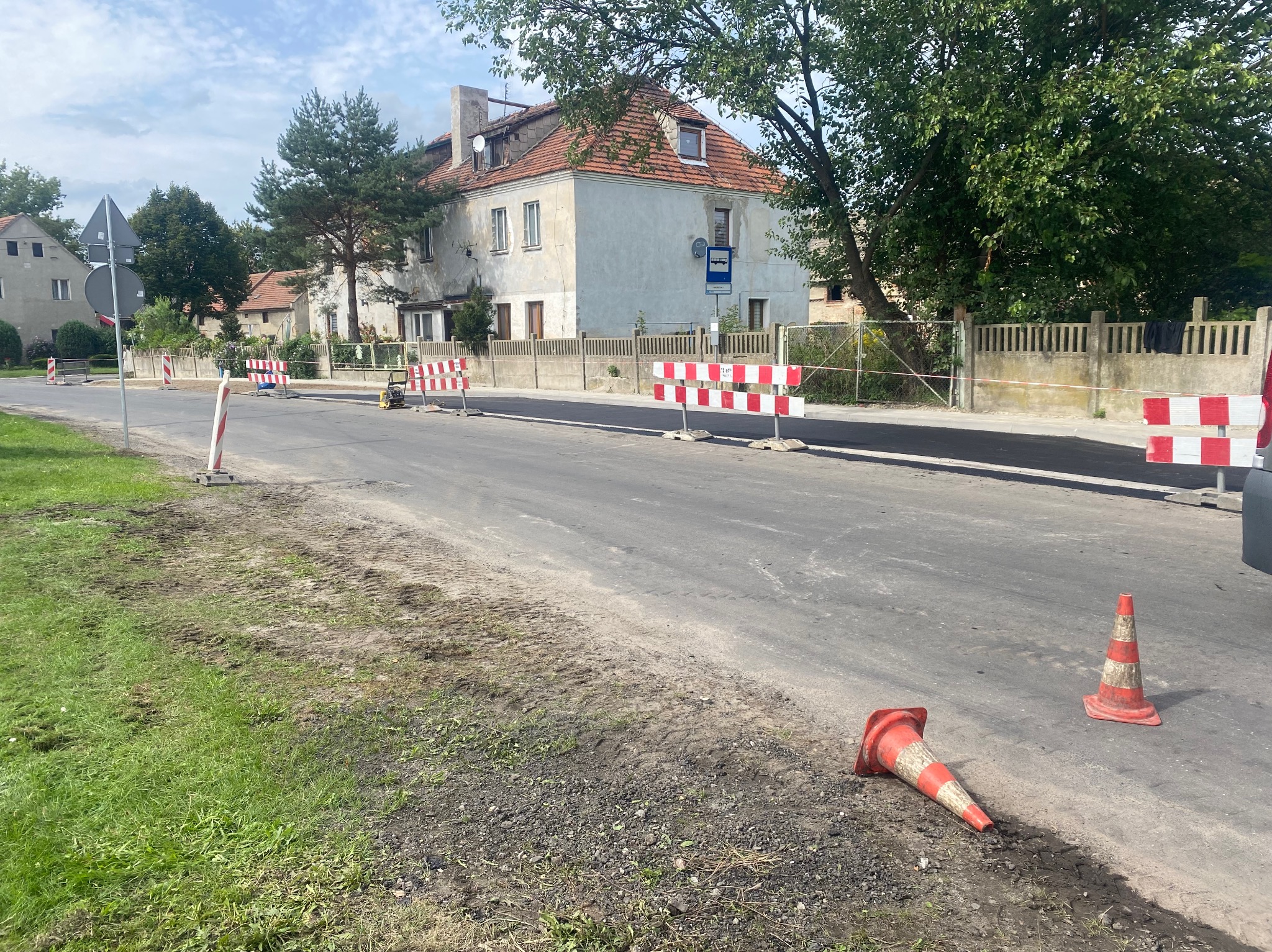 Świeżo położony asfalt na drodze w obszarze zabudowanym, zabezpieczony barierkami i pachołkami, z jednym pachołkiem leżącym na poboczu. W tle budynki mieszkalne i drzewa.