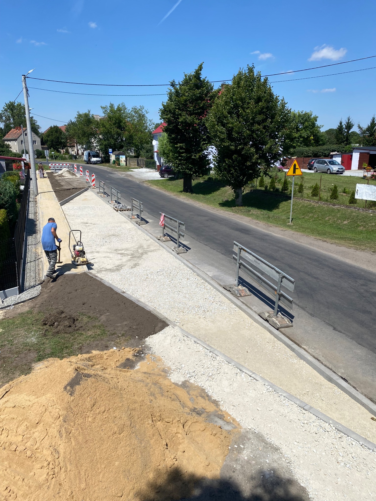 Pracownik w niebieskiej koszulce zagęszcza piasek ubijarką wibracyjną podczas budowy chodnika z kostki, widoczny hałda piasku, obrzeża chodnika i ulica w tle.