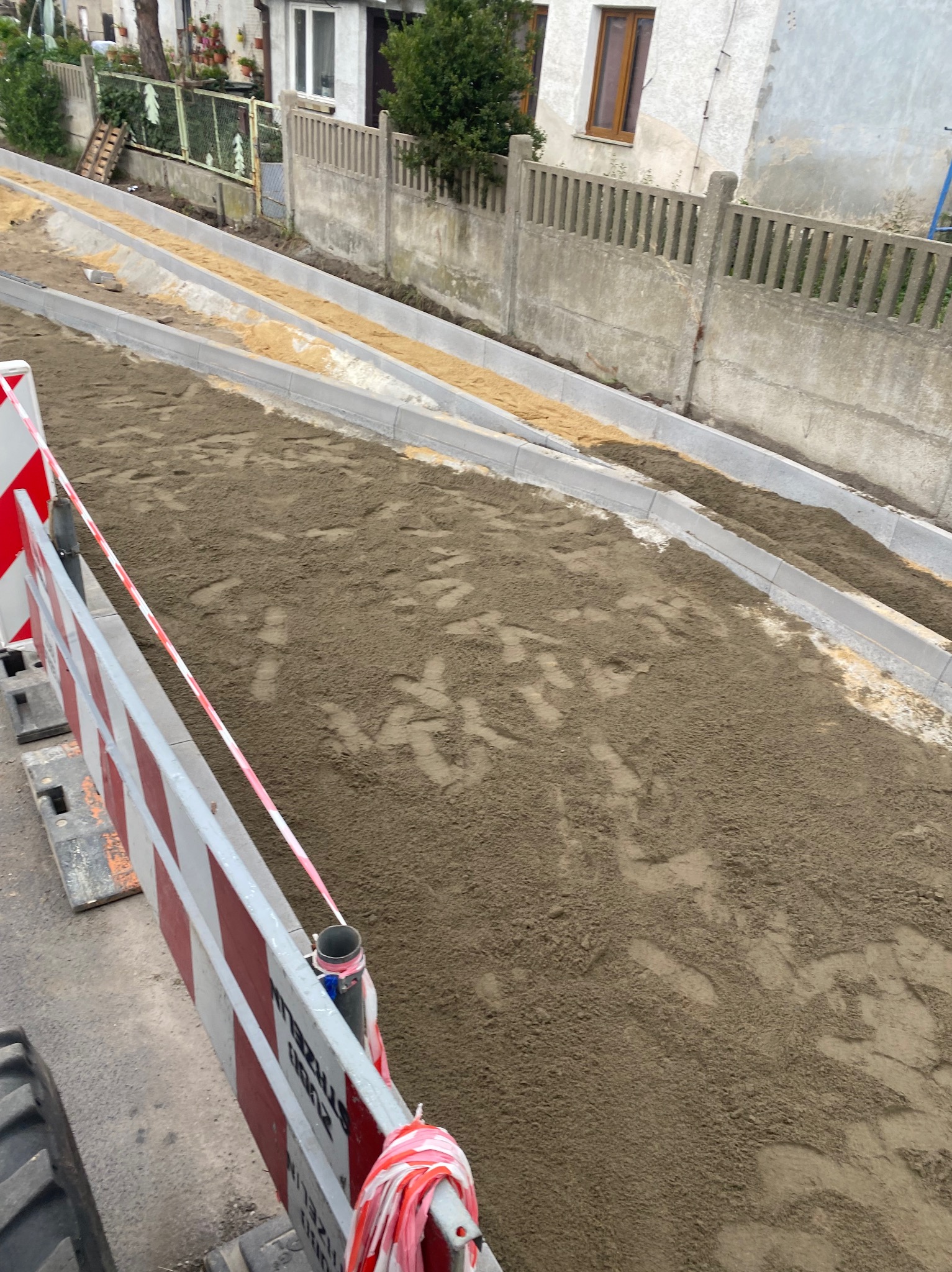 Przygotowanie podłoża pod kostkę brukową: wyrównany teren z piaskiem i obrzeżami, widoczne zabezpieczenia budowlane w postaci barierek.