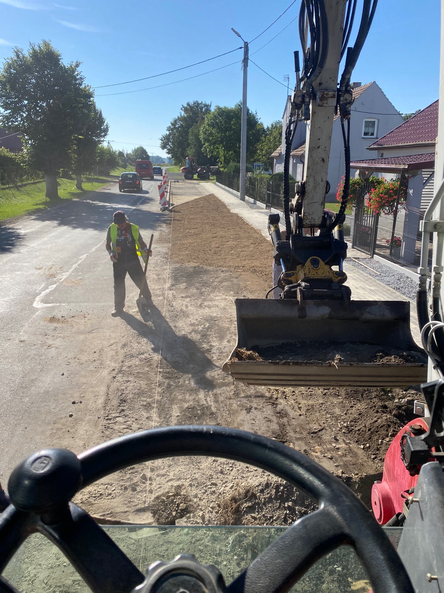 Widok z kabiny koparki na pracownika z łopatą przygotowującego podłoże pod chodnik z kostki brukowej. W tle ulica z samochodami i domy.