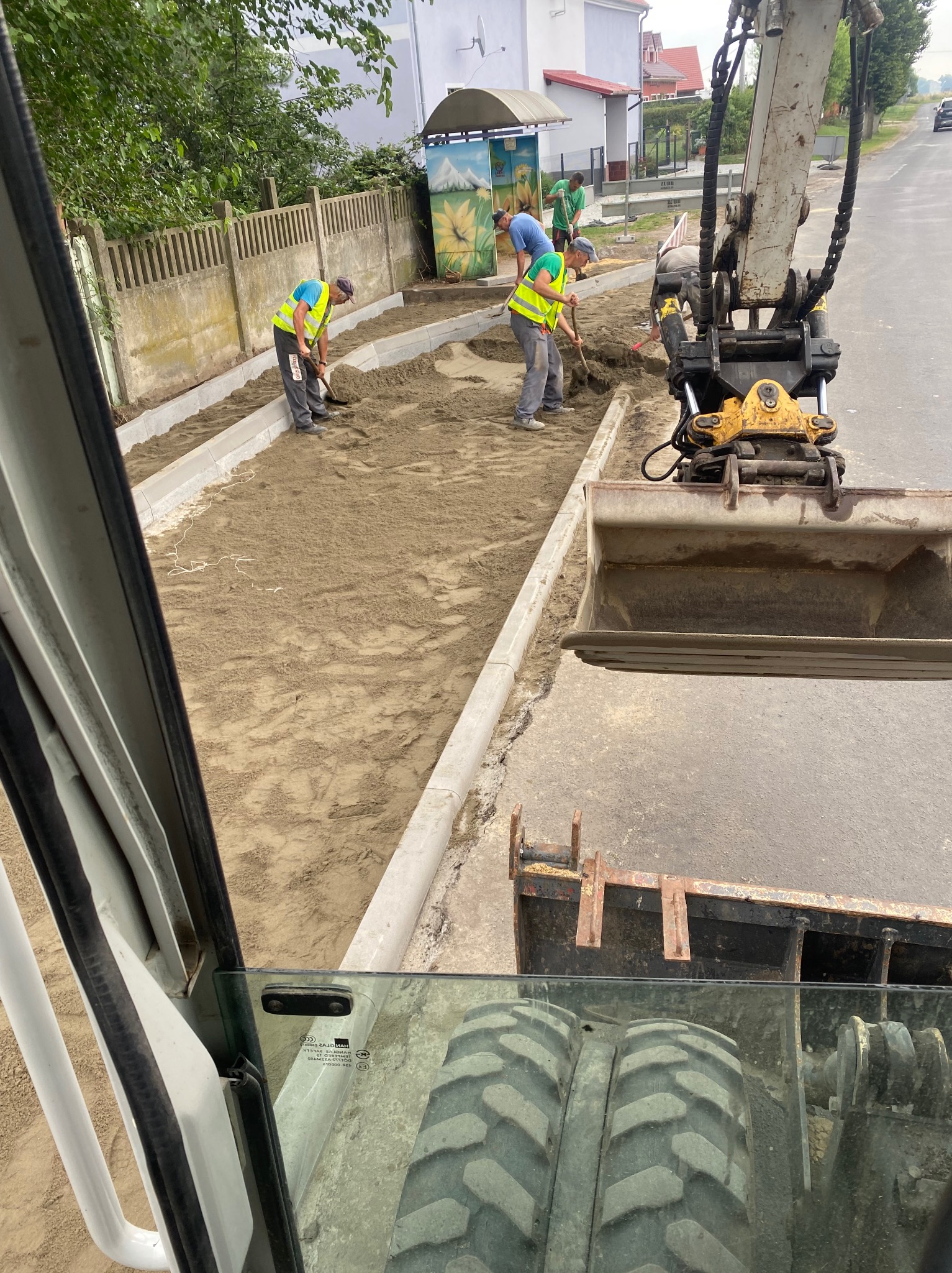 Widok z kabiny koparki na robotników wyrównujących piasek pod chodnik, widoczne krawężniki i budynek w tle.