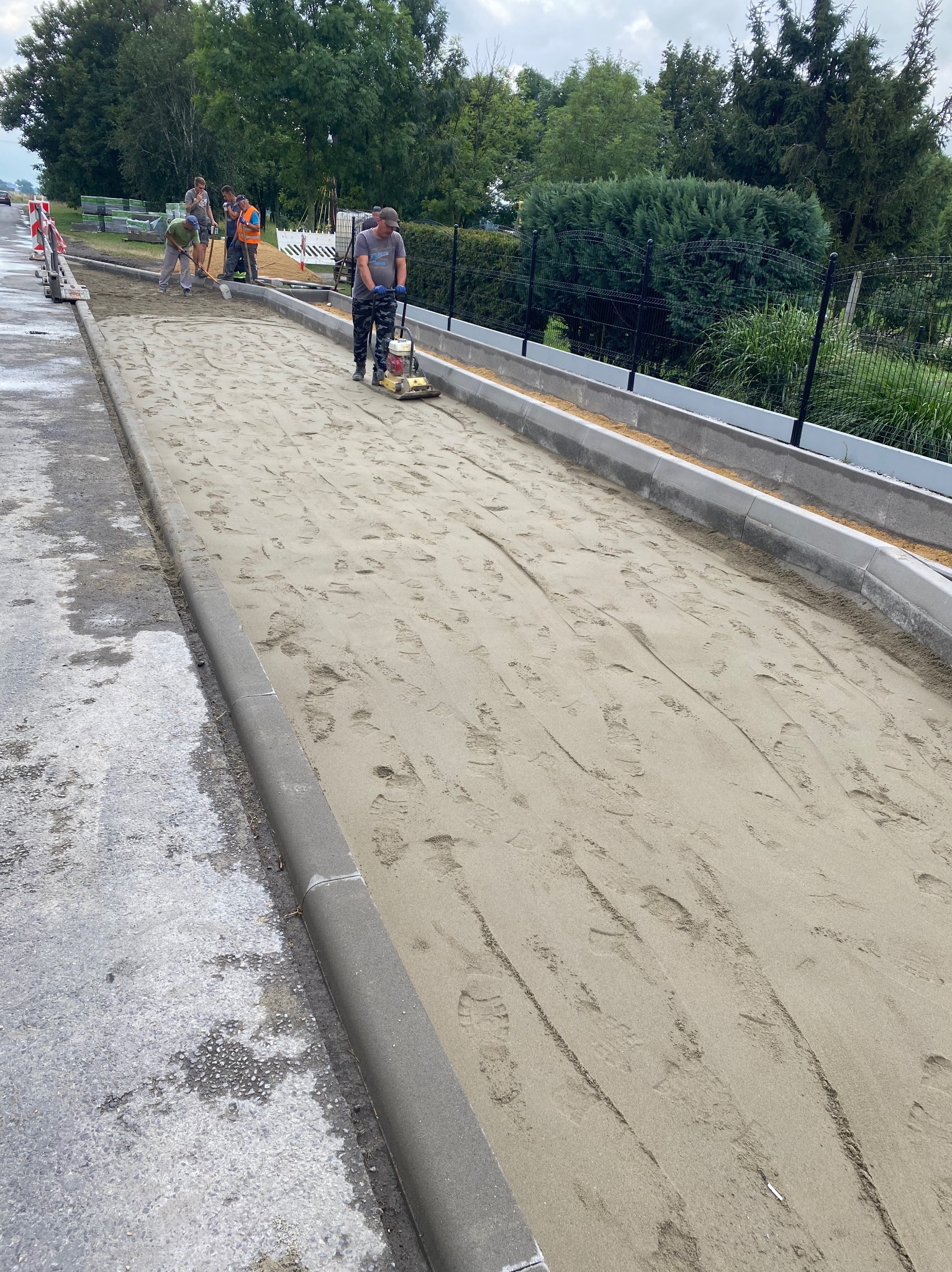 Robotnicy przygotowują podłoże pod chodnik z kostki, ubijając piasek zagęszczarką wibracyjną, widoczne nowe krawężniki, w tle drzewa.