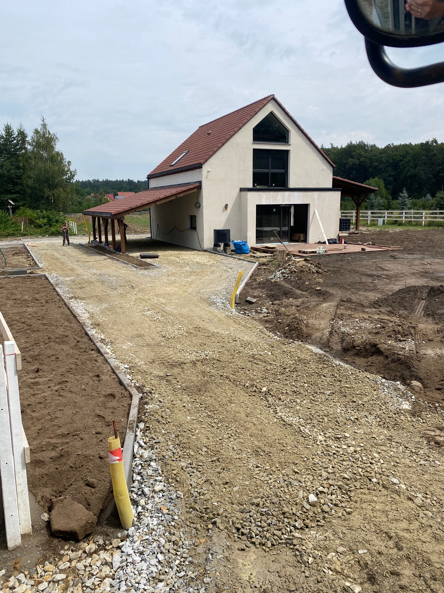 Przygotowanie podłoża z kruszywa pod przyszły podjazd do nowoczesnego domu z drewnianą wiatą, widoczna ziemia ogrodowa, elementy instalacji podziemnej i pracownik w oddali.
