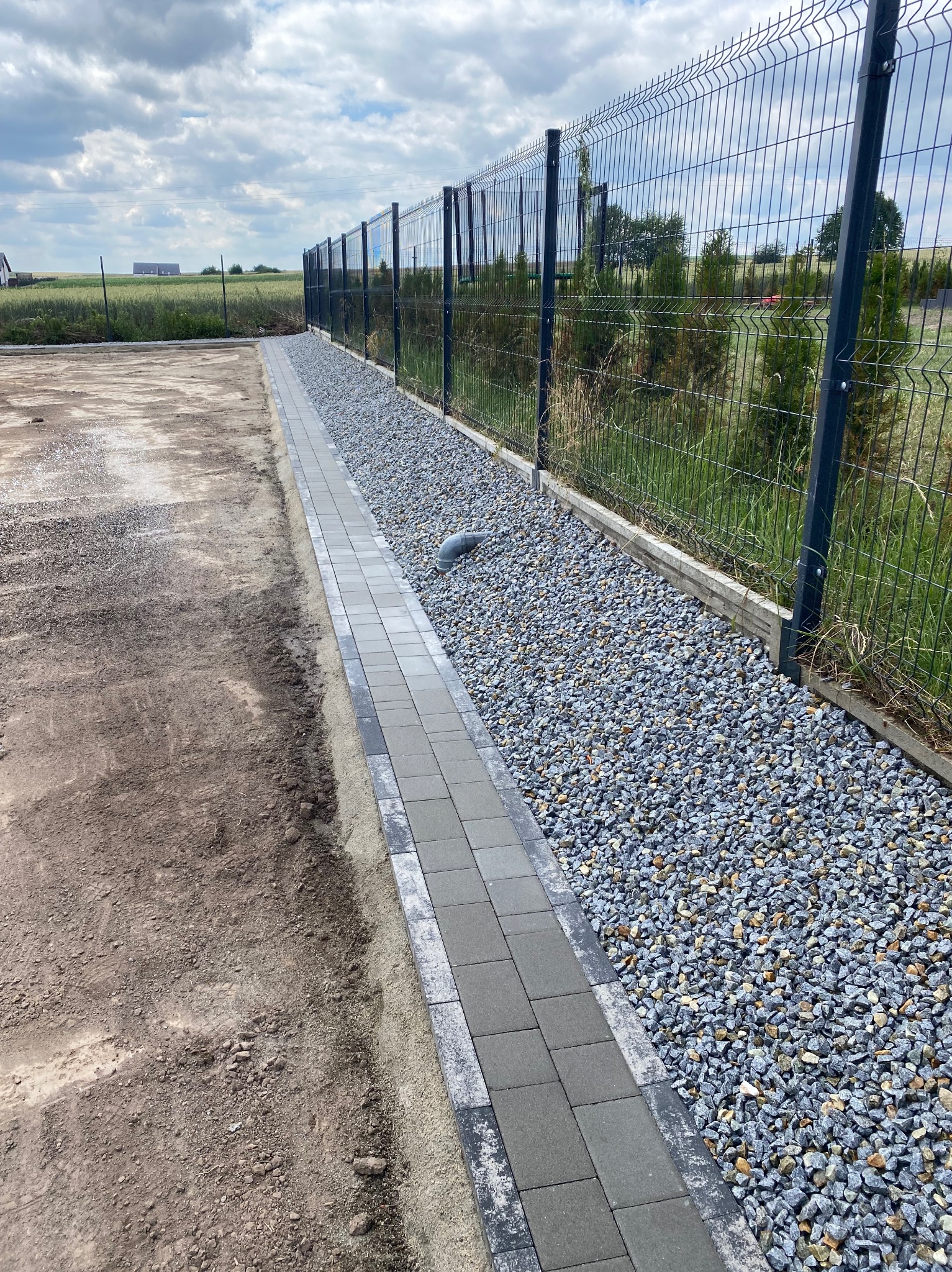Świeżo ułożony chodnik z szarych kostek brukowych obok metalowej siatki ogrodzeniowej i wysypanego żwiru, z widoczną rurą kanalizacyjną i przygotowanym terenem pod dalsze prace.