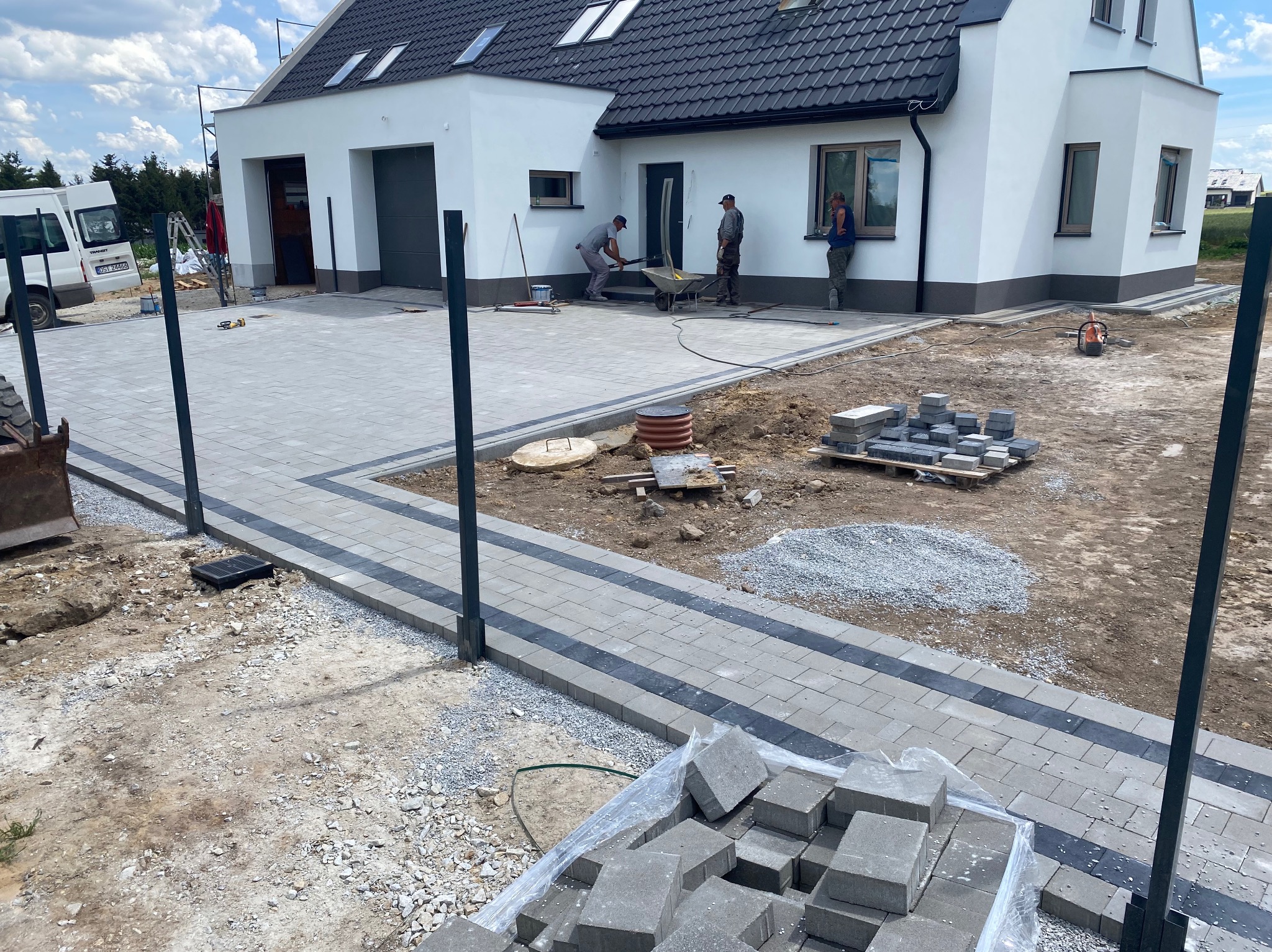 Układanie kostki brukowej na posesji domu jednorodzinnego, widoczni pracownicy, taczka, paleta z kostką i częściowo ułożony chodnik z szarej kostki z czarnymi wstawkami.