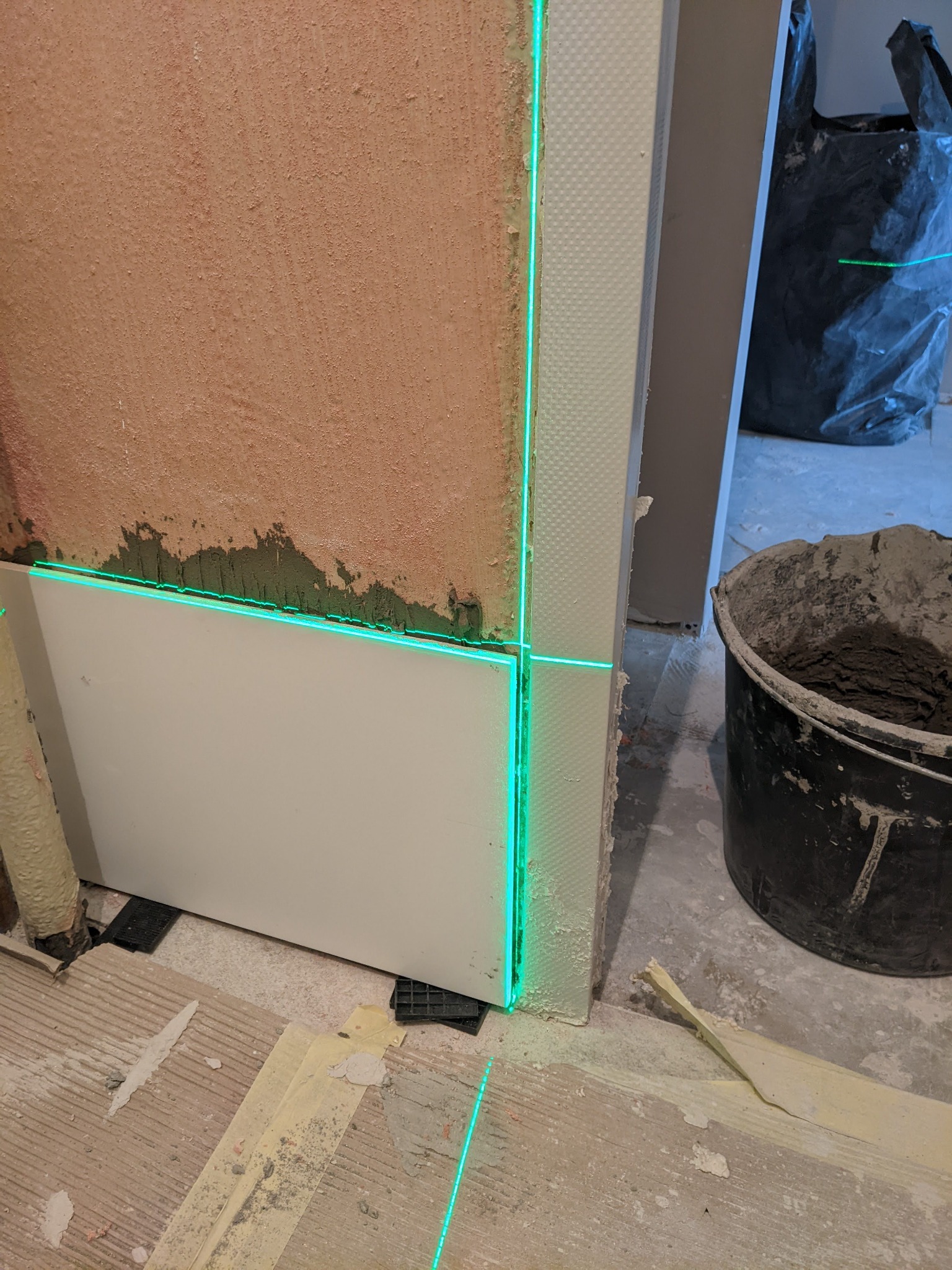 Fragment ściany z nałożoną zaprawą i białymi panelami, wyznaczony poziom za pomocą lasera krzyżowego, widoczny kubeł z zaprawą i resztki materiałów na podłodze.