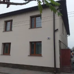 Dwupiętrowy dom z jasną elewacją, brązowymi ramami okiennymi i kamienną podmurówką, widoczne tabliczka z numerem domu, fragment drewnianej bramy i pracownik z taczką w tle.