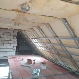 Poddasze w trakcie ocieplania wełną mineralną, widoczne metalowe profile konstrukcyjne, biała cegła, narzędzia pracy i kartony na podłodze z desek.