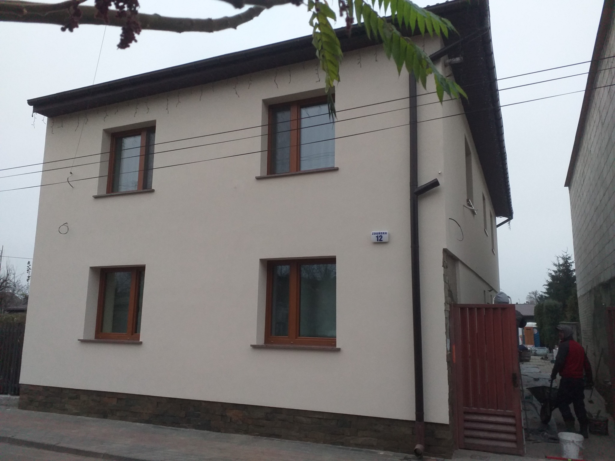 Dwupiętrowy dom z jasną elewacją, brązowymi ramami okiennymi i kamienną podmurówką, widoczne tabliczka z numerem domu, fragment drewnianej bramy i pracownik z taczką w tle.