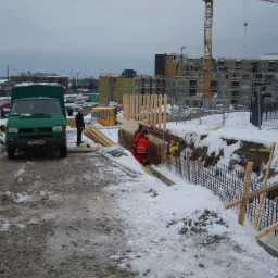 Budowa fundamentów budynku w zimowych warunkach: robotnicy w pomarańczowych kombinezonach pracują przy zbrojeniu, obok zaparkowany zielony bus, w tle budynek w trakcie budowy i żuraw.