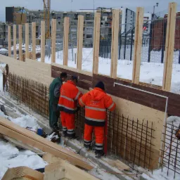 Trzech pracowników w odblaskowych kombinezonach montuje zbrojenie do szalunku drewnianego na zasypanym śniegiem placu budowy, widoczne nowe bloki mieszkalne w tle.