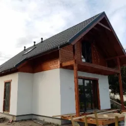 Niedokończony dom szkieletowy z drewnianą elewacją na górze i białą na dole, czarny dach z trzema kominami, widoczne rusztowanie i elementy konstrukcyjne werandy.