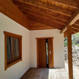 Wykończony drewniany podbitka dachowa i stolarka okienna w nowym domu z białymi ścianami, widok na wejście i fragment tarasu z desek.