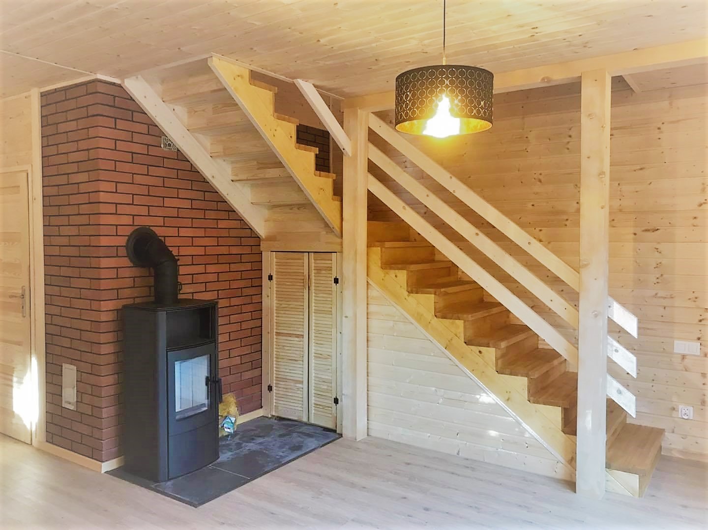 Wnętrze domu z drewnianą boazerią, schodami z jasnego drewna, kominkiem na tle ceglanej ściany i lampą wiszącą z ażurowym abażurem.