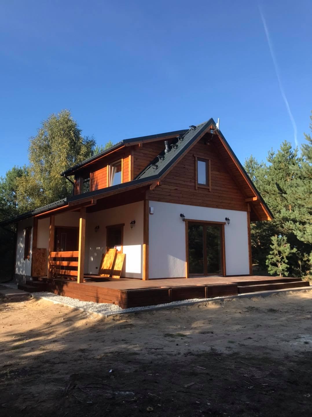 Dwukondygnacyjny dom z drewnianą elewacją na piętrze i białym tynkiem na parterze, z zadaszonym tarasem i ciemnym dachem, otoczony drzewami.