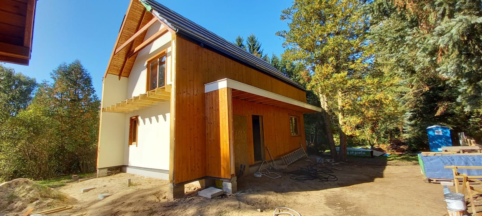 Surowy stan dwukondygnacyjnego domu z drewnianą elewacją i częściowo otynkowanymi ścianami, widoczna konstrukcja dachu i fundamenty na betonowych bloczkach, otoczenie w stanie budowy z widocznymi...