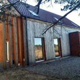 Nowoczesny dom z drewnianą elewacją i ceglanymi elementami, widoczny dach z oknami połaciowymi. Ujęcie z zewnątrz, w naturalnym otoczeniu.